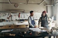 Two craftsmen discussing plans in a well-equipped workshop with tools and blueprints.